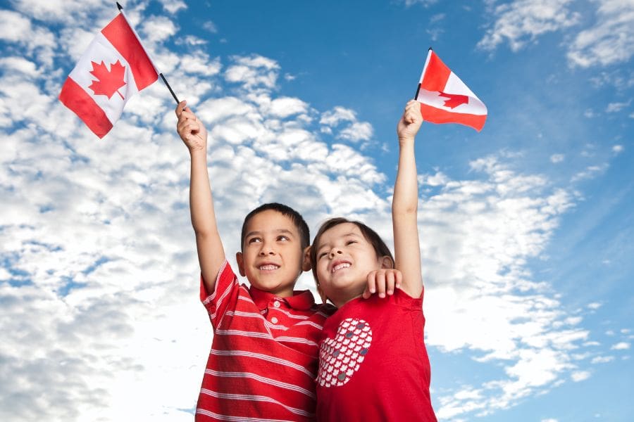 Two young smiling children wearing red shirts and holding small Canadian flags high in the air against a blue sky with light clouds.