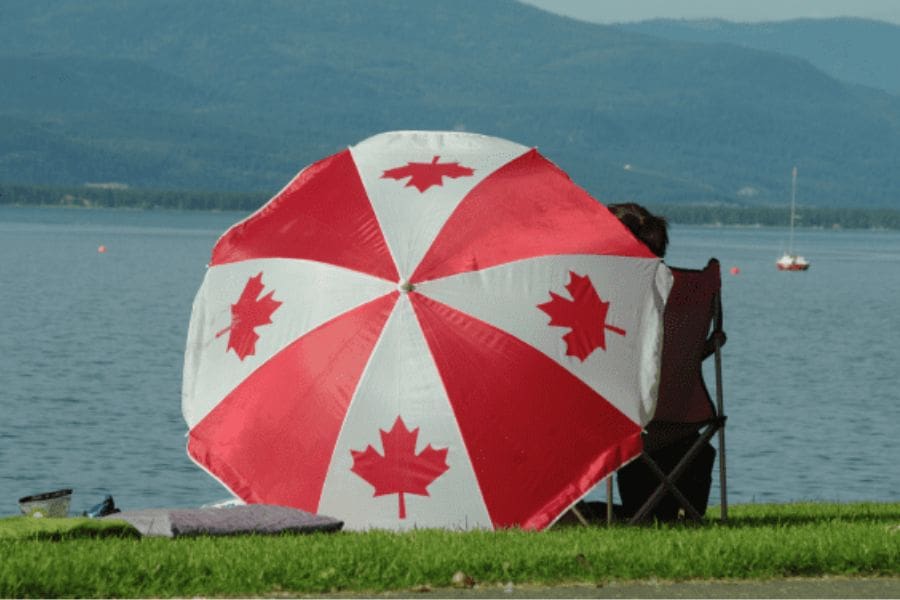 A person relaxing in a lawn chair next to a large red and white maple leaf umbrella on a grassy lakeside shore with a small sailboat in the distance.