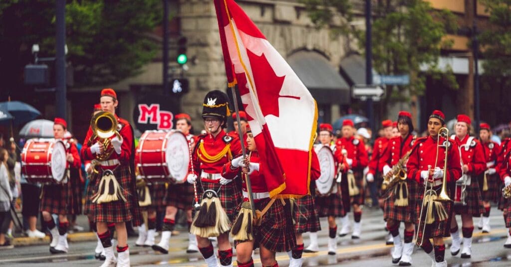 A marching band in red uniforms parading down a city street while waving a large Canadian flag during a Canada Day celebration.