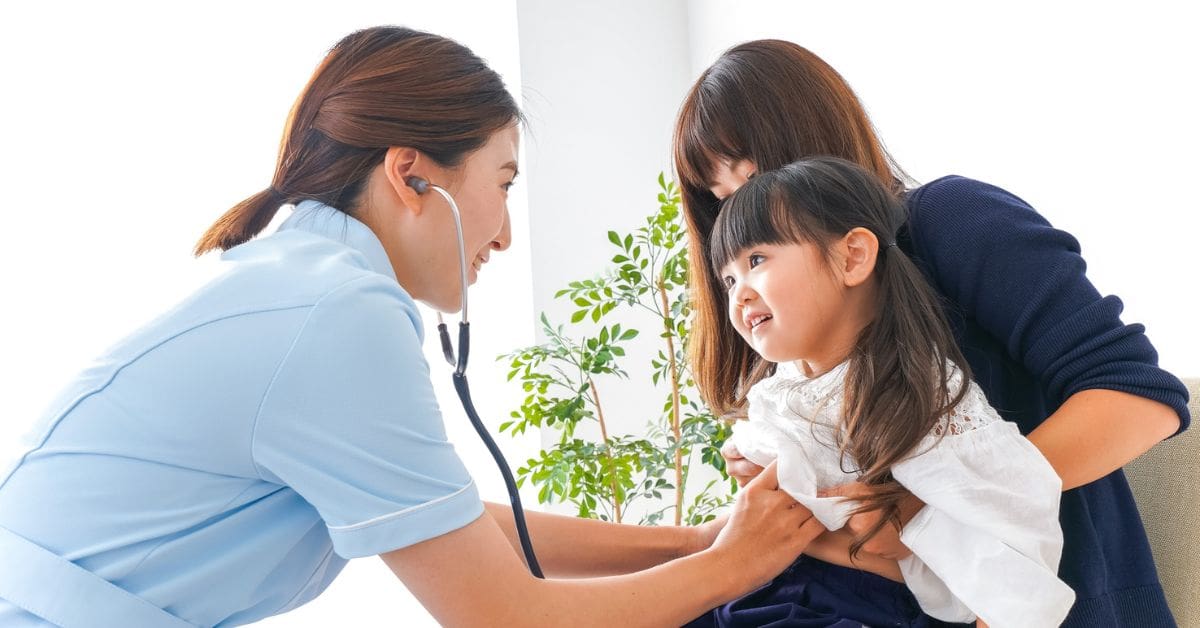 A friendly female family doctor checking a young girl's breathing with a stethoscope while her smiling mother holds her during a checkup.