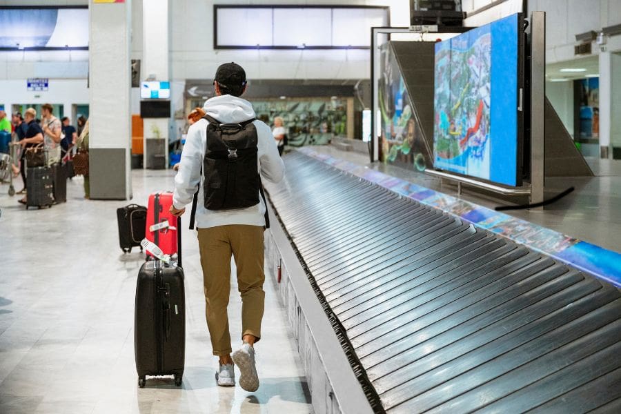 A male traveller walking through a bright airport baggage claim area while pulling a suitcase and wearing a backpack after landing.