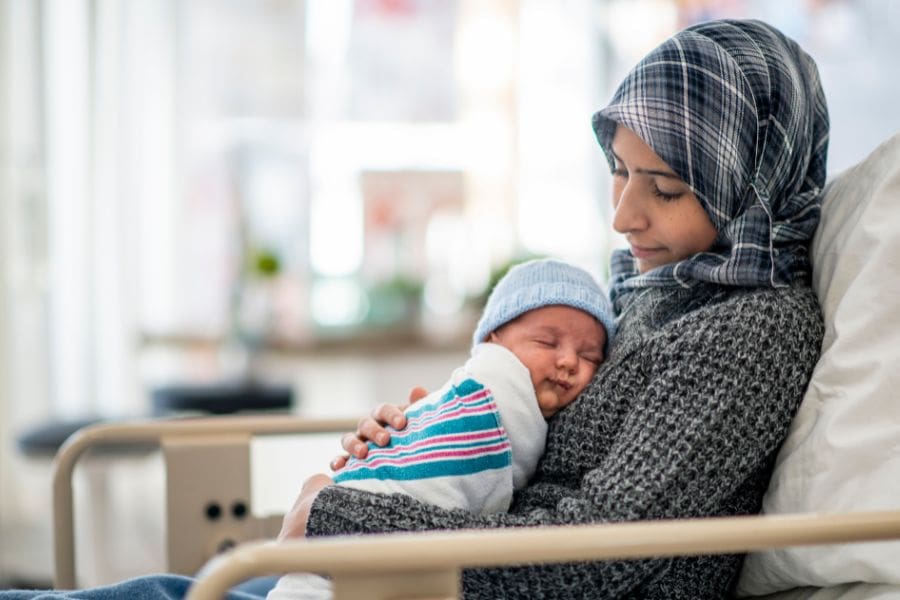 A smiling mother wearing a hijab holds her swaddled newborn baby close while resting in a hospital bed after giving birth.