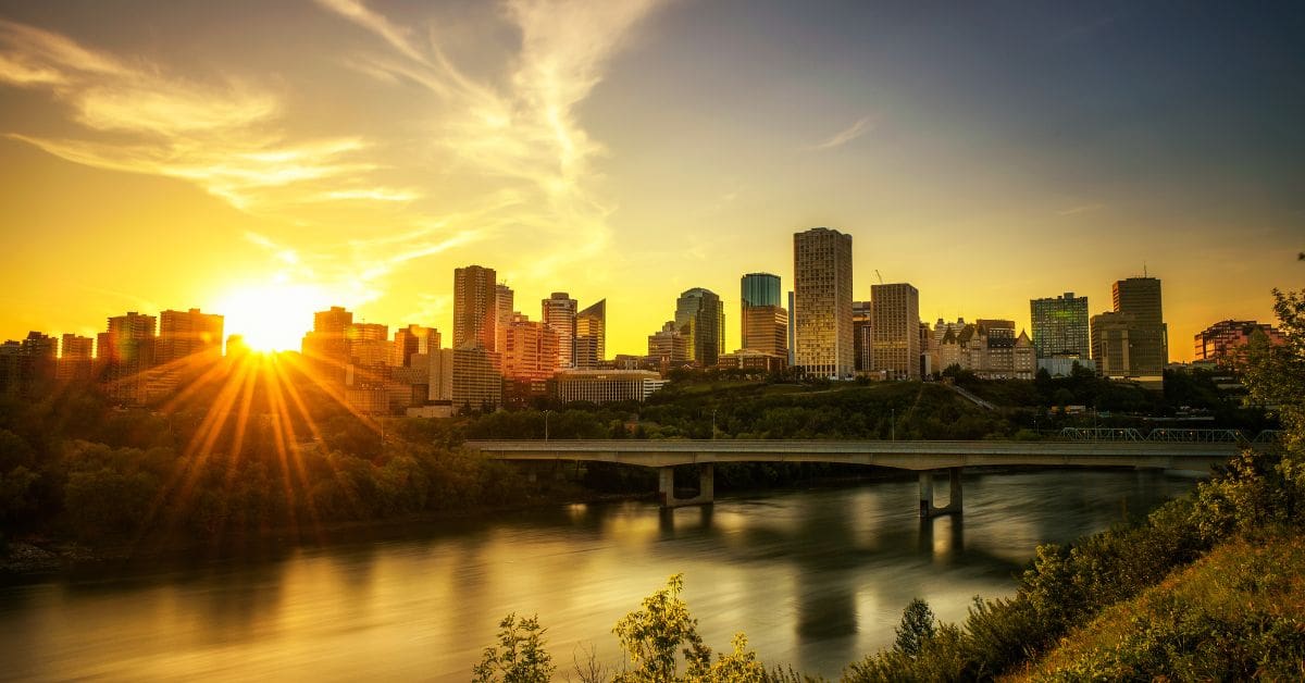The downtown Edmonton skyline at sunset, with a brilliant sunburst reflecting over the river and green valley trees.