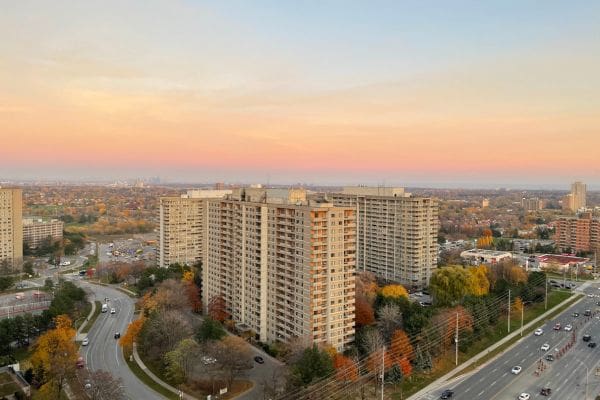Aerial view of high-rise apartment buildings surrounded by autumn trees. The Toronto skyline and Lake Ontario are on the horizon.