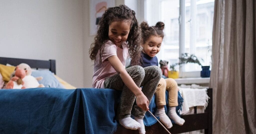 Two young sisters smiling and playing together while sitting on a bed in a shared room.