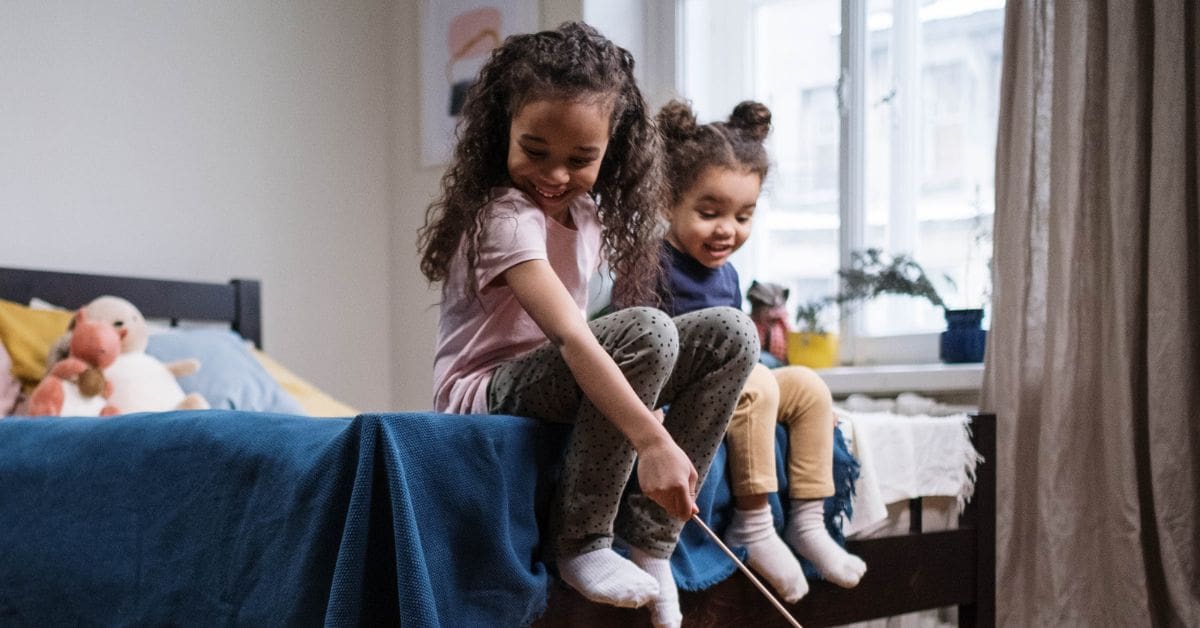 Two young sisters smiling and playing together while sitting on a bed in a shared room.