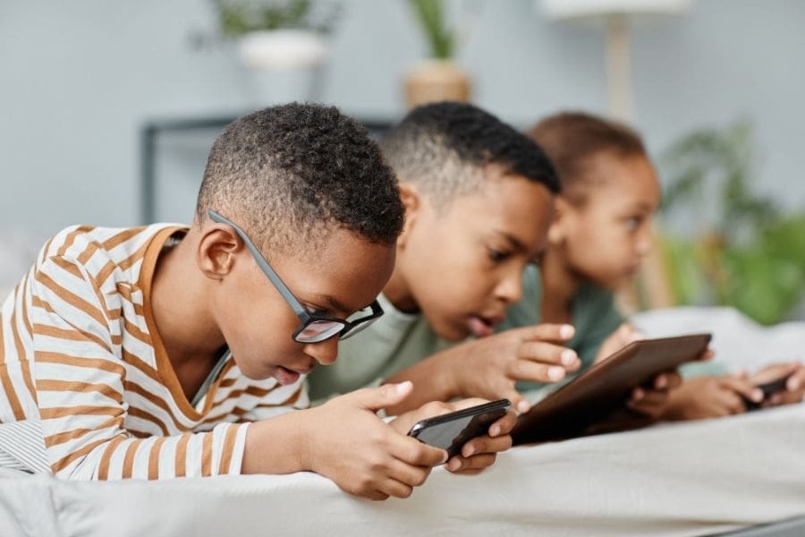 Three young siblings lying side-by-side on a bed, focused on their phones and tablets in a shared bedroom. CMHC's National Occupancy Standards can help you decide how many bedrooms you need.