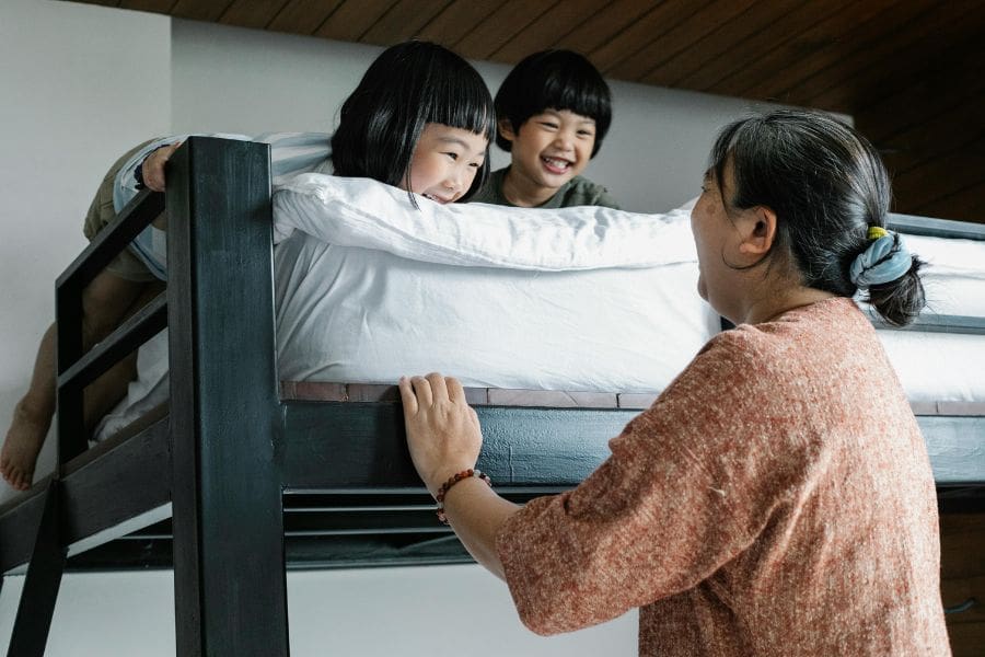 A smiling grandmother looking up at her two grandchildren playing on the top bunk of a black wooden bunk bed in their bedroom.