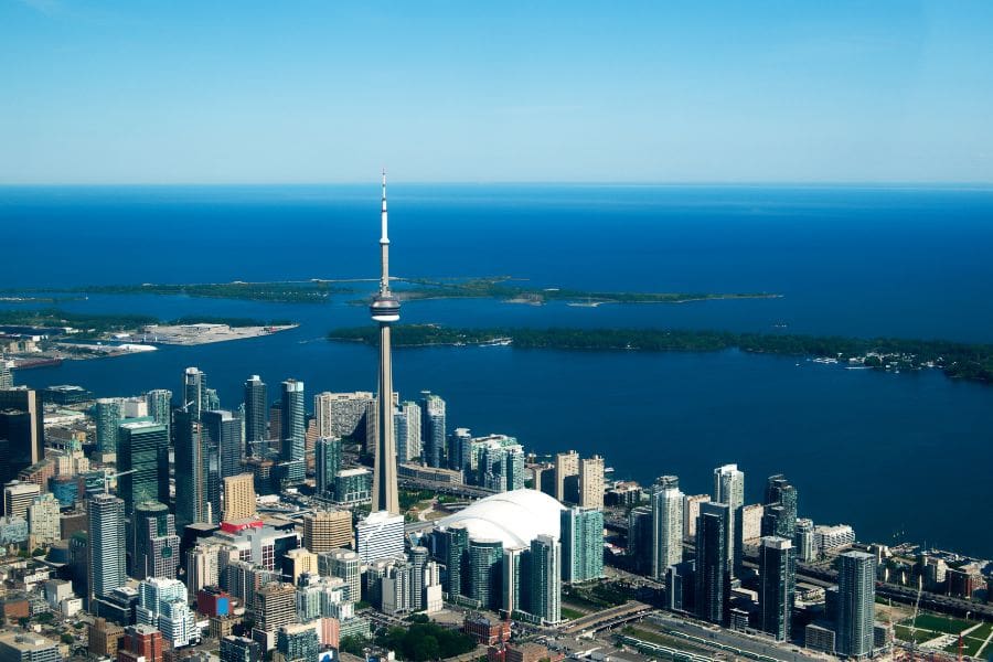 Aerial, high-angle view of downtown Toronto showing the CN Tower, high-rise office and condo buildings, the Rogers Centre dome, the Toronto Islands, and the deep blue water of Lake Ontario on a clear day.
