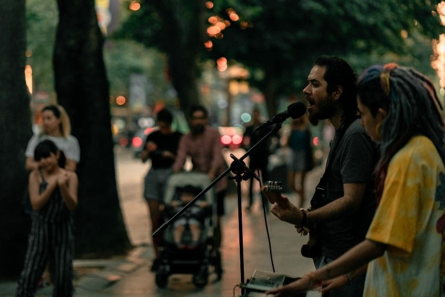 Street musicians singing and playing guitar outdoors on a tree-lined city sidewalk at a summer busker festival while a crowd watches.