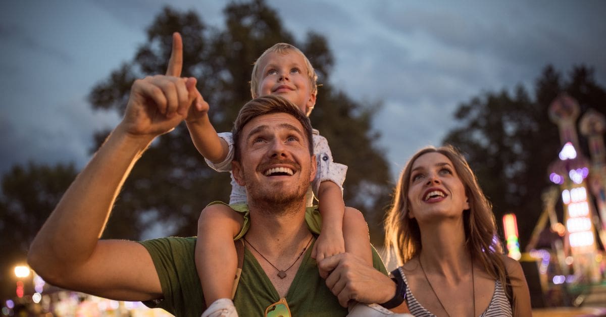 A happy family with a young child on his father's shoulders looking up at carnival rides and lights during an outdoor summer festival in Canada.