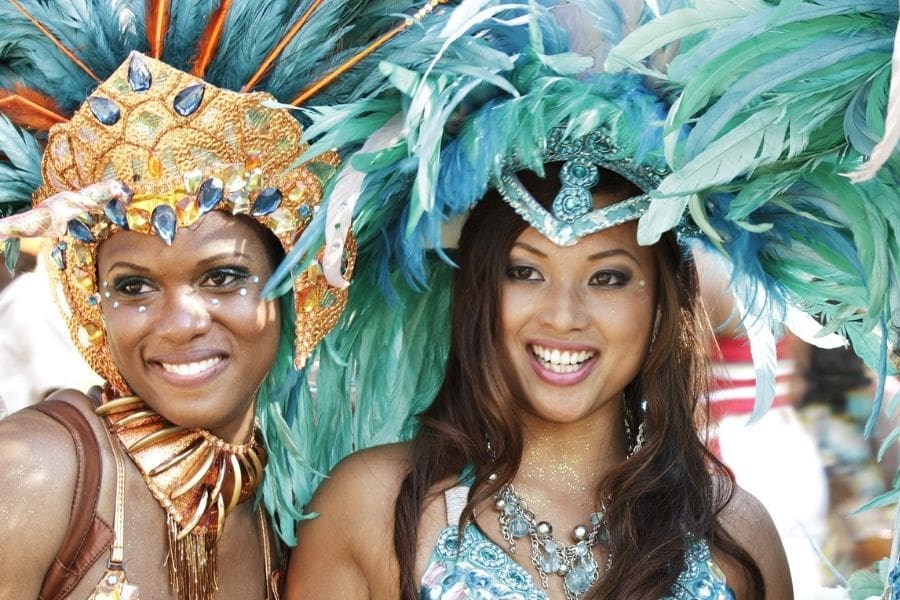 Two smiling women wearing colorful feather headpieces and jeweled Caribbean Carnival costumes at a summer festival in Canada.