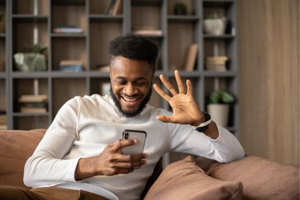 A smiling newcomer sitting on a couch waving during a smartphone video call using a Canadian SIM card.