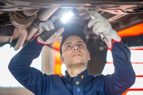 A licensed mechanic in a workshop inspects the underbody of a used car with an inspection light.