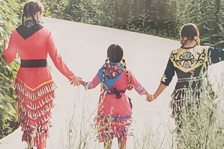 Three Indigenous children wearing traditional ribbon dresses and jingle dresses walking hand in hand down a path.