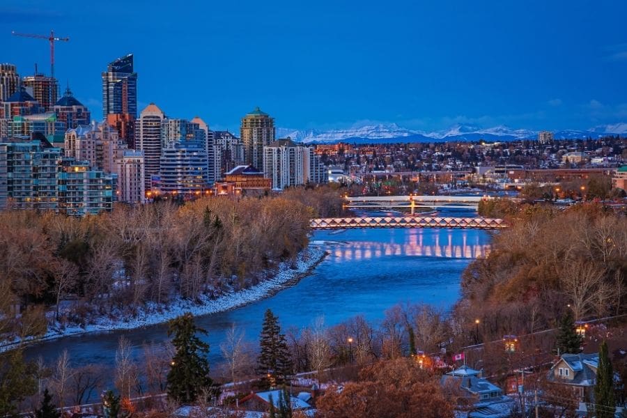 The red Peace Bridge spanning the Bow River is lit up at  dusk with high-rise residential apartments and the Rocky Mountains in the background, representing the best neighbourhoods in Calgary.