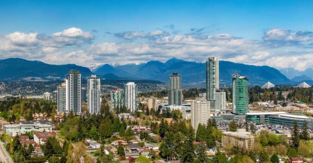 Surrey BC skyline with high-rise residential towers, lush green trees, and snow-capped mountains in the background.