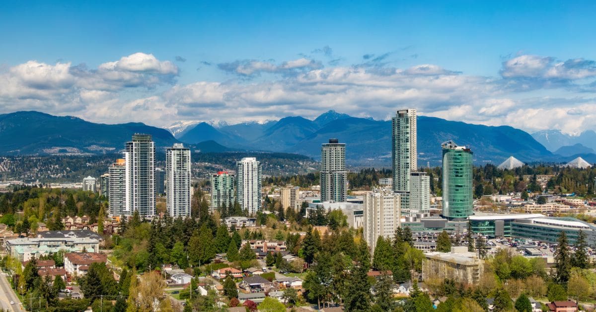 Surrey BC skyline with high-rise residential towers, lush green trees, and snow-capped mountains in the background.