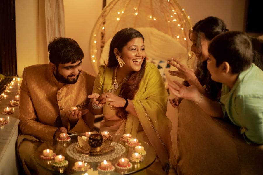 Adults and children in traditional Indian attire sitting together around a table filled with lit oil lamps and tea light diyas